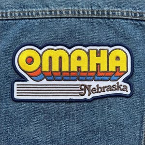 May include: An embroidered patch featuring the word OMAHA in large yellow letters with orange and blue drop shadows, positioned above the word Nebraska in brown script and five horizontal brown lines, all on a blue denim fabric background.