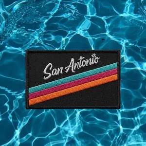 May include: A rectangular black embroidered patch featuring the white cursive text San Antonio. Below the text are three diagonal stripes in teal, magenta, and orange that run from the bottom left corner toward the right side of the patch.