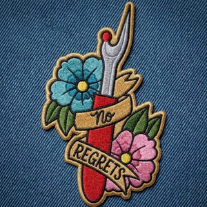 May include: An embroidered patch featuring a red seam ripper with a silver tip, surrounded by a blue flower and a pink flower with green leaves. A tan ribbon banner wraps around the tool with the black text No REGRETS written across it.