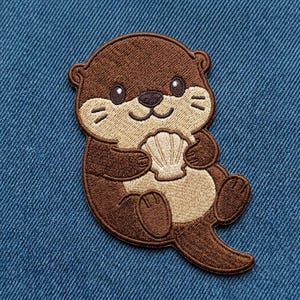 May include: An embroidered patch featuring a brown otter with a light tan belly and face, holding a small, light-colored seashell. The otter has dark eyes, a small nose, and whiskers, with a thick brown tail extending to the lower right side.