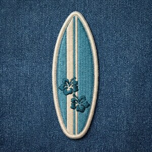 May include: An embroidered patch shaped like a surfboard with a pointed top and rounded bottom. The design features a teal blue background with a vertical cream-colored stripe down the center and two dark teal hibiscus flowers on the lower half.