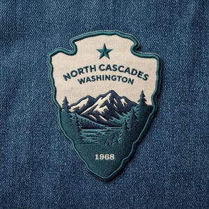 May include: A shield-shaped embroidered patch featuring a cream-colored background with dark teal borders. It displays a mountain range, evergreen trees, a star, and the text NORTH CASCADES WASHINGTON 1968 in dark teal thread on denim fabric.