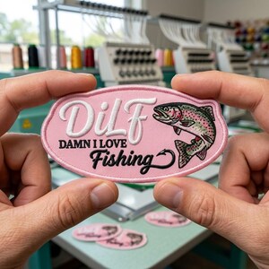 May include: An oval-shaped embroidered patch with a light pink background. It features the white text "DiLF" and black text "DAMN I LOVE Fishing" with a fish hook integrated into the word Fishing. A colorful trout is embroidered on the right side.