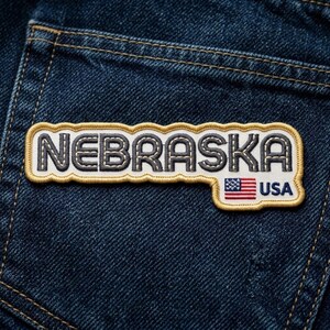 May include: A rectangular embroidered patch with a scalloped gold border on a dark blue denim fabric. The patch features the word NEBRASKA in dark blue block letters on a white background, with a small American flag and the text USA below it.