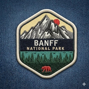 May include: A hexagonal embroidered patch featuring a mountain range with a red sun against a pale yellow sky. A dark blue band displays the white text BANFF NATIONAL PARK above a green forest scene with a small red bear silhouette at the bottom.