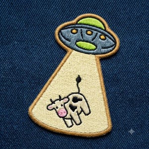 May include: An embroidered patch featuring a gray UFO with a bright green dome and yellow lights. A light yellow beam of light extends downward from the craft, containing a small white cow with black spots and a pink nose, all outlined in brown.