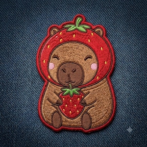 May include: An embroidered patch featuring a brown capybara wearing a red strawberry-shaped hood with green leaves. The capybara has pink cheeks, a smiling expression, and is holding a small red strawberry with green leaves in its front paws.