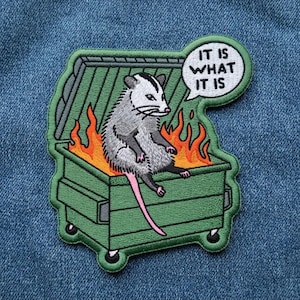 May include: An embroidered patch featuring a gray and white opossum sitting inside a green dumpster filled with orange and yellow flames. A white speech bubble above the opossum contains the black text IT IS WHAT IT IS in capital letters.