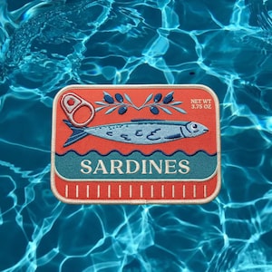 May include: A rectangular embroidered patch featuring a coral orange background with a light blue sardine, a silver pull tab, and a dark blue olive branch. The text SARDINES and NET WT 3.75 OZ are embroidered in white on the front of the design.
