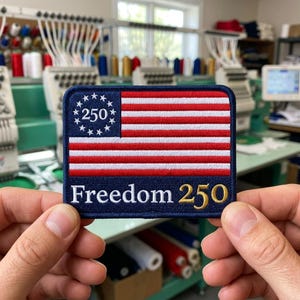 May include: A rectangular embroidered patch featuring a stylized American flag with a blue canton containing a circle of white stars around the number 250. Below the flag, the text Freedom 250 is embroidered in white and gold thread.