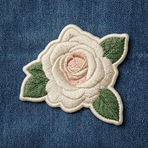 May include: An embroidered patch featuring a cream-colored rose with a soft pink center and three dark green leaves. The floral design is stitched with visible thread texture and is positioned against a dark blue denim fabric background.