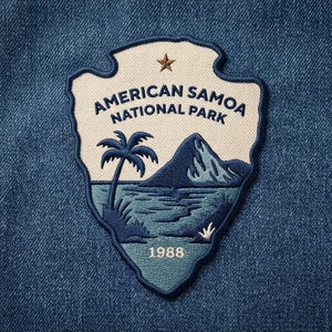 May include: An embroidered patch shaped like a shield featuring the text AMERICAN SAMOA NATIONAL PARK and 1988. The design includes a brown star, a palm tree, a mountain, and ocean waves in shades of blue and cream on a dark blue denim background.