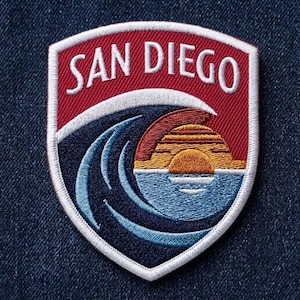 May include: A shield-shaped embroidered patch featuring the text SAN DIEGO in white letters on a red background. Below the text, a dark blue ocean wave curls over a stylized orange and yellow sunset reflecting on light blue water.