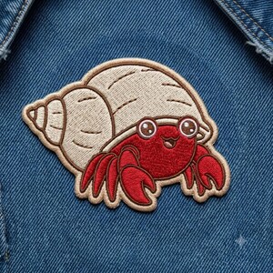 May include: An embroidered patch featuring a red hermit crab with large eyes and a smiling mouth, emerging from a beige spiral seashell. The patch is stitched with detailed thread work and has a tan border, displayed on blue denim fabric.