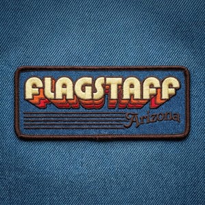 May include: A rectangular embroidered patch featuring the word FLAGSTAFF in large, cream-colored block letters with orange and red drop shadows. Below the text, the word Arizona is written in a script font, accompanied by five horizontal lines.