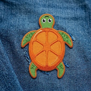 May include: An embroidered patch shaped like a turtle with a bright orange shell resembling a citrus slice and a green head and flippers. The turtle has a small, curved line on its head and is attached to a textured blue denim fabric surface.
