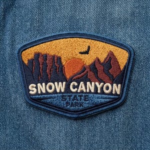 May include: An embroidered patch on blue denim fabric featuring a landscape of dark blue and brown mountains against a yellow sky with a setting sun and a bird silhouette. The text reads SNOW CANYON STATE PARK in white capital letters.