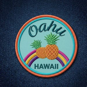 May include: A circular embroidered patch with a light blue background, featuring two yellow pineapples above a colorful rainbow. The text "Oahu" is embroidered in dark green at the top, and "HAWAII" is embroidered in dark green at the bottom.