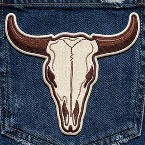 May include: An embroidered patch featuring a cream-colored steer skull with long, curved brown horns. The skull has dark brown accents around the eye sockets and nasal cavity, with a thick cream border, attached to a dark blue denim fabric background.
