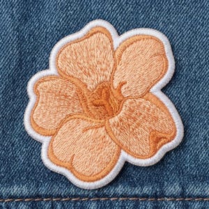 May include: An embroidered patch in the shape of a five-petaled flower. The flower features shades of light orange and peach thread with a darker orange center, all outlined by a thick white border against a dark blue denim fabric background.