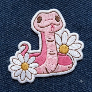 May include: An embroidered patch featuring a pink snake with a light pink belly, dark brown eyes, and a small pink tongue. The snake is coiled and surrounded by two white daisies with yellow centers, all outlined in a thick white border.