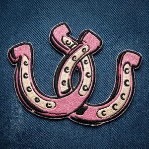 May include: An embroidered patch featuring two overlapping horseshoe shapes. The horseshoes are colored in shades of light pink and cream, with black outlines and black circular nail hole details throughout the design, set against a blue denim fabric.