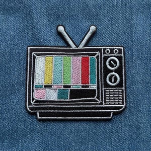 May include: An embroidered patch shaped like a vintage television set with two antennas. The screen displays a colorful test pattern with vertical stripes in yellow, blue, green, pink, and red, alongside a black and white lower section.