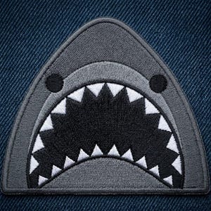 May include: An embroidered patch shaped like a shark head, featuring a dark gray upper section, a lighter gray snout area, two small black circular eyes, and a wide open mouth filled with sharp white triangular teeth against a black background.
