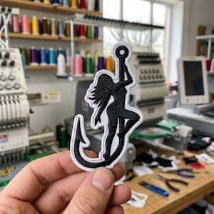 May include: A black and white embroidered patch featuring a silhouette of a figure climbing a large fishing hook. The design is rendered in solid black thread with a thick white border, creating a high-contrast graphic suitable for fabric application.