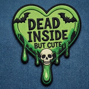 May include: A heart-shaped embroidered patch featuring a lime green slime design with black borders. The text reads DEAD INSIDE BUT CUTE in black letters. The patch includes two black bats at the top and a beige skull at the bottom center.