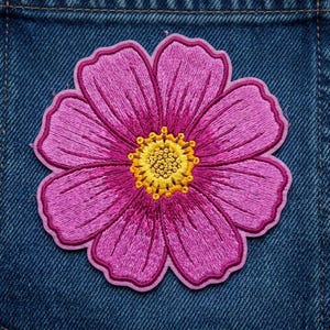 May include: A circular embroidered patch featuring a vibrant pink flower with eight distinct petals. The center of the flower is composed of dense yellow and orange thread work, creating a textured, circular stamen detail against the pink petals.