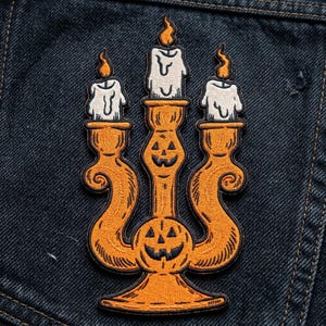 May include: An embroidered patch featuring an orange candelabra with two jack-o'-lantern faces. The candelabra holds three white candles with orange flames, each showing dripping wax details. The design is outlined in black thread on a dark denim fabric.