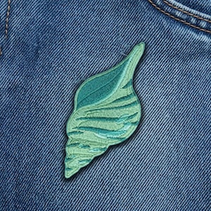 May include: An embroidered patch in the shape of a seashell, featuring various shades of green thread. The design displays a spiral shell structure with detailed stitching that creates a textured appearance, set against a blue denim fabric background.