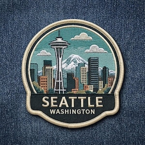 May include: A circular embroidered patch featuring the Seattle skyline, the Space Needle, and a snow-capped mountain against a teal sky with white clouds. The bottom black section displays the white text SEATTLE WASHINGTON with a cream border.