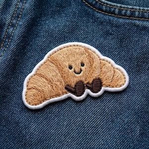 May include: An embroidered patch shaped like a tan croissant with a smiling face and two small dark brown arms. The patch features a white stitched border and is attached to a piece of dark blue denim fabric with visible stitching details.