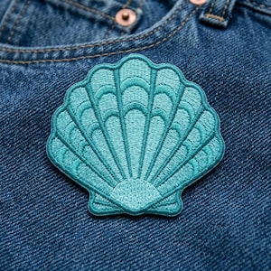 May include: A teal embroidered patch in the shape of a scallop shell with detailed radiating ridges and a fan-like pattern. The patch is stitched with thread in varying shades of blue-green, creating a textured surface on a dark blue denim fabric.
