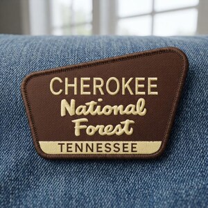 May include: A brown embroidered patch with a trapezoidal shape featuring the text CHEROKEE National Forest in cream-colored lettering, with the word TENNESSEE printed in a cream-colored rectangular bar at the bottom of the patch.