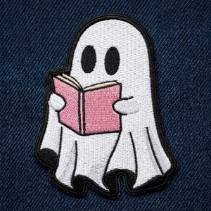 May include: An embroidered patch featuring a white ghost with black oval eyes, holding an open pink book. The ghost has a black border and black accents at the bottom of its sheet-like form, with the entire design stitched onto a dark blue denim fabric.