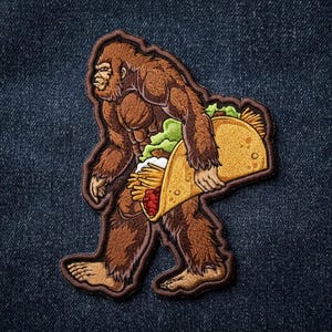 May include: An embroidered patch featuring a brown, furry Bigfoot creature walking while holding a large yellow taco filled with green lettuce, thin yellow fries, and red meat, all outlined with a dark brown border on a dark blue denim fabric.