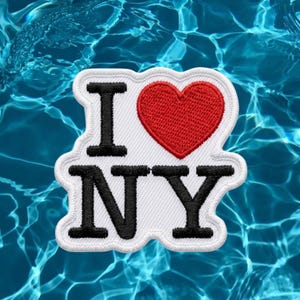 May include: A white embroidered patch featuring the black text I and NY with a red heart symbol in the center. The patch has a white border and a die-cut shape that follows the contour of the letters and the heart, creating a clean, bold design.