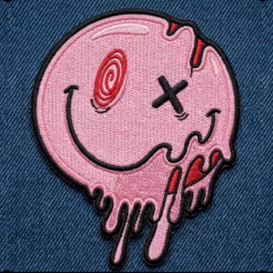 May include: A circular embroidered patch featuring a light pink melting smiley face. The design includes a red spiral eye, a black X-shaped eye, and a wide black curved mouth. The bottom edge of the face drips downward with black outlines.