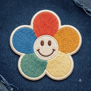 May include: A flower-shaped embroidered patch featuring a central cream-colored smiley face with brown eyes and a curved mouth. The five petals are stitched in vibrant red, orange, yellow, green, and blue thread, all outlined in a cream border.