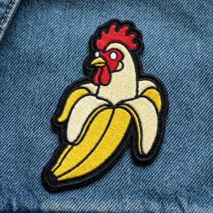 May include: An embroidered patch featuring a white chicken head with a red comb and wattle emerging from a peeled yellow banana. The design is outlined in thick black thread and is attached to a blue denim fabric background with visible texture.