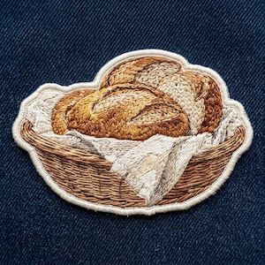 May include: An embroidered patch featuring a woven brown basket filled with golden-brown loaves of bread. The bread rests on a white cloth liner with visible folds. The entire design is outlined with a cream-colored border on a dark blue background.