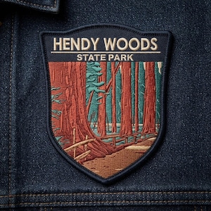 May include: A shield-shaped embroidered patch on dark blue denim fabric. The patch features a forest scene with tall reddish-brown trees and teal foliage. The text HENDY WOODS STATE PARK is stitched in white at the top of the patch.