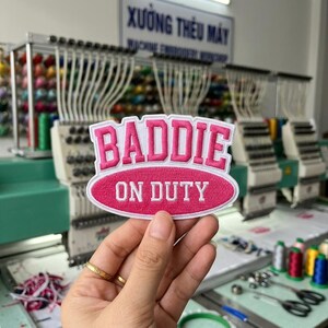 May include: An oval-shaped embroidered patch featuring the word BADDIE in large pink letters above the text ON DUTY in white letters, all set against a pink background with a white border, held up in front of industrial embroidery machines.