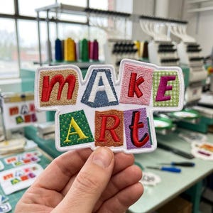 May include: A rectangular embroidered patch featuring the words MAKE ART in colorful, textured block letters. Each letter is set against a unique fabric-like background pattern, including tan, blue denim, pink lace, green gingham, green polka dot, and purple.