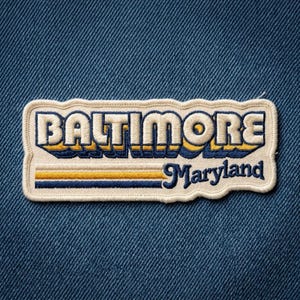 May include: A rectangular embroidered patch with a cream-colored border featuring the word BALTIMORE in large, bold, dark blue letters with a yellow and blue shadow effect, and the word Maryland in script below with yellow and blue stripes.