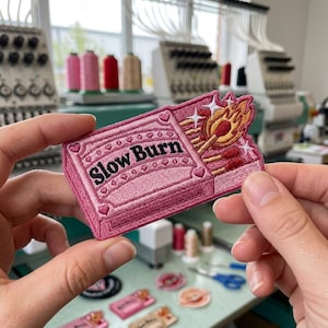 May include: An embroidered patch shaped like a pink matchbox with the text Slow Burn in black letters. The design features a lit match with orange and yellow flames, several unlit matches, and small white star accents against a pink background.