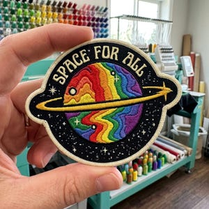 May include: A circular embroidered patch featuring a black background with a central planet displaying wavy rainbow-colored stripes. A yellow ring encircles the planet, and the text "SPACE FOR ALL" is stitched in gold above the planet.
